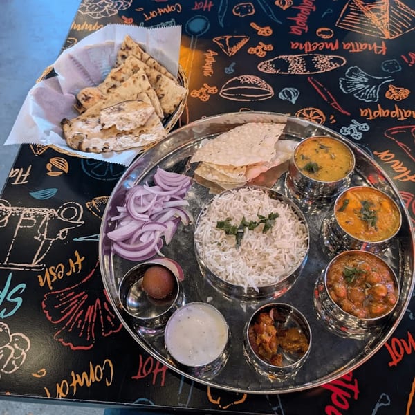 A Bansee's Kitchen thali with rice, curries, naan, papadum, onions, and dessert on a patterned table.